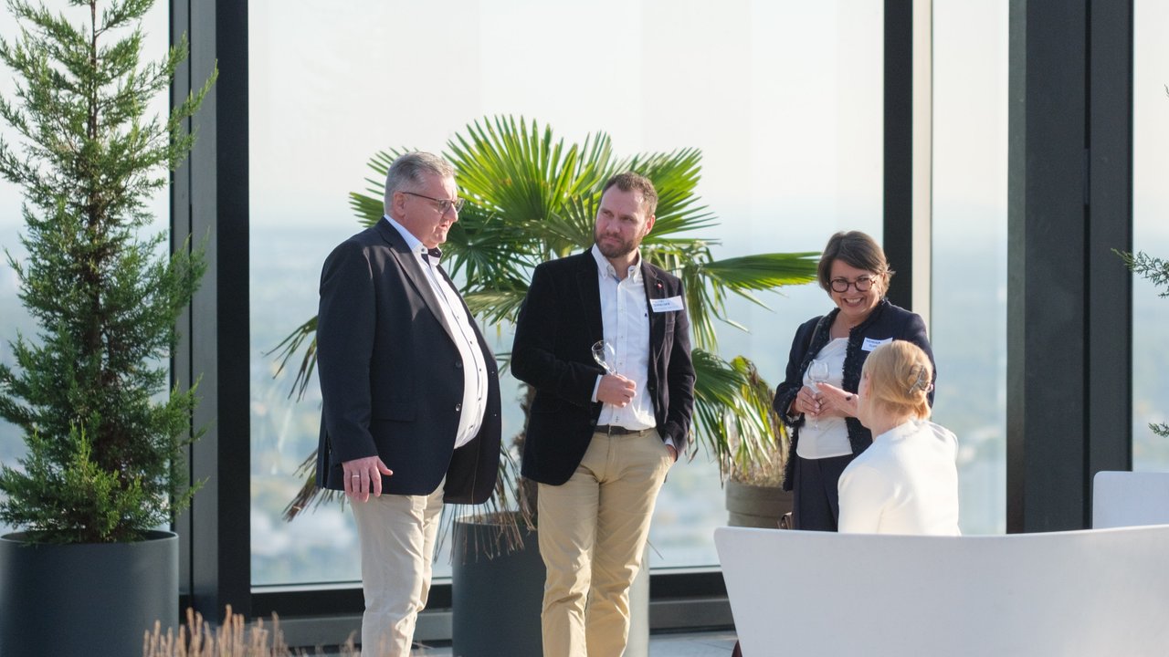 Four people converse indoors with large windows and plants in the background, one holding a glass.