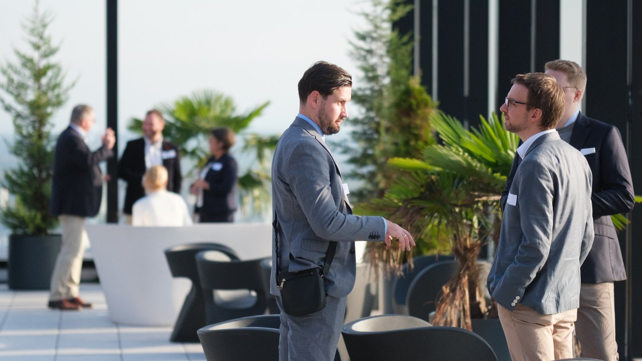 Two men in suits converse on a rooftop with palm trees and other people networking in the background.