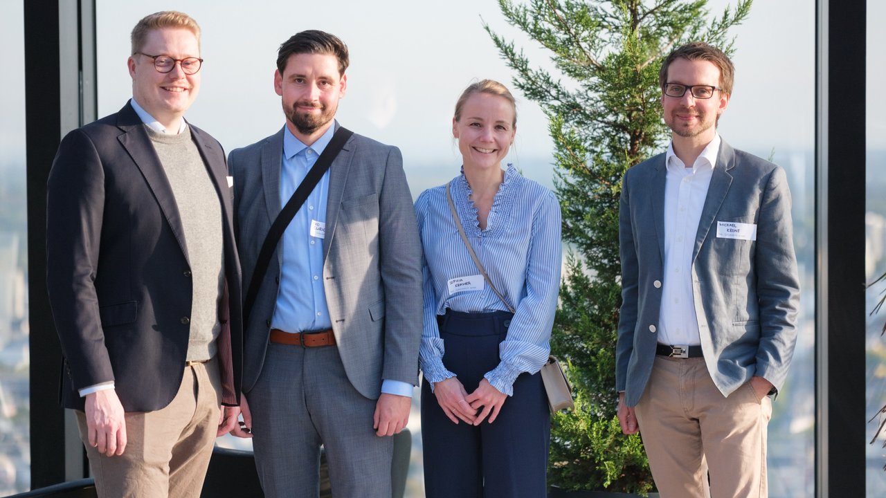 Four people stand together in a modern setting with a plant in the background, smiling at the camera.