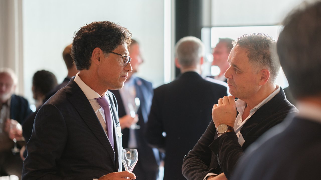 Two men in business attire engage in conversation at a social event, with other attendees in the background.