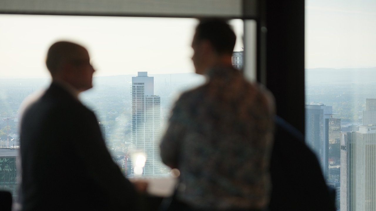 Two blurred figures stand by a window overlooking a city skyline with tall buildings in the background.