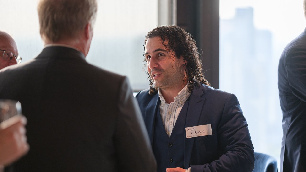 A man with curly hair in a suit talks to another man, with a city view in the background.