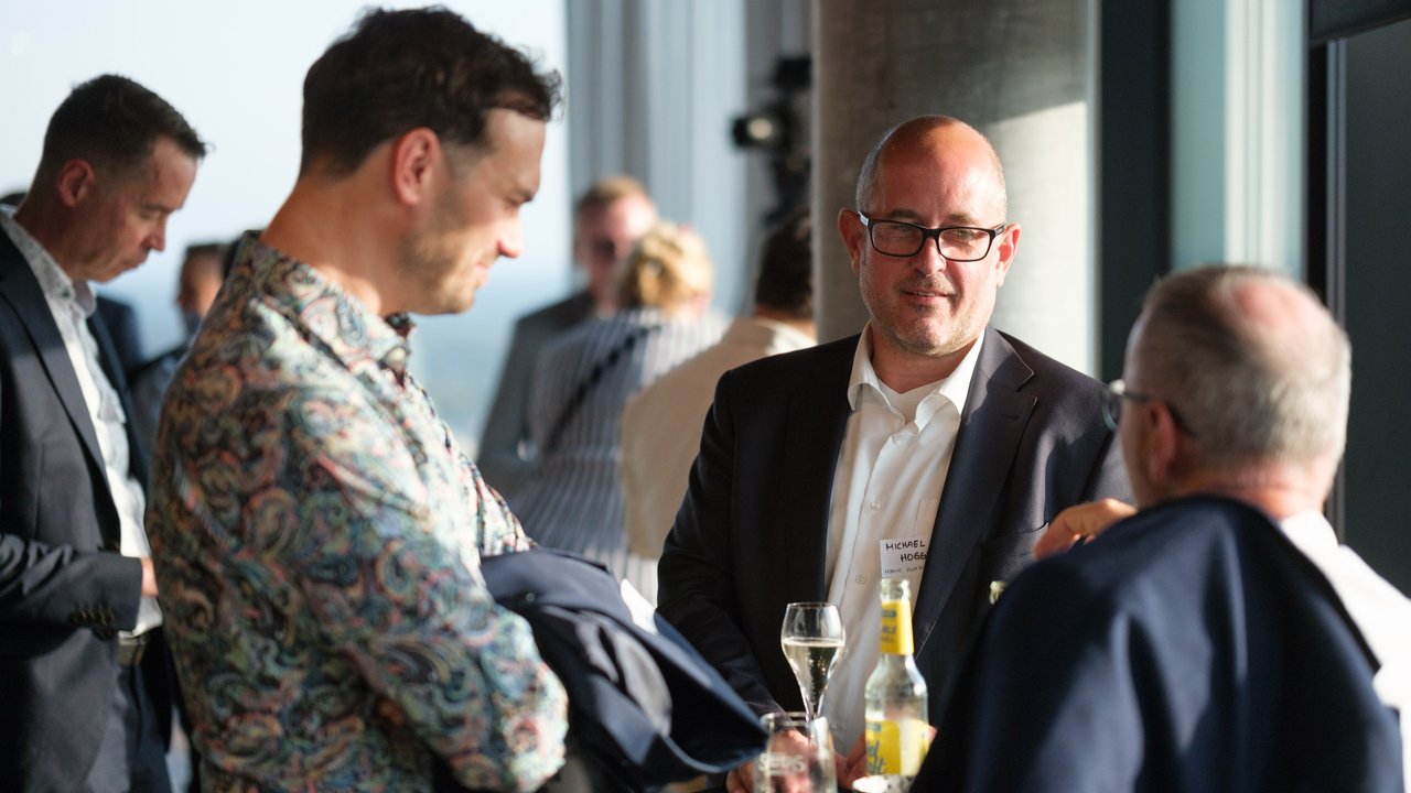 A group of men in business attire conversing at a social event, with drinks on a table and a city view in the background.