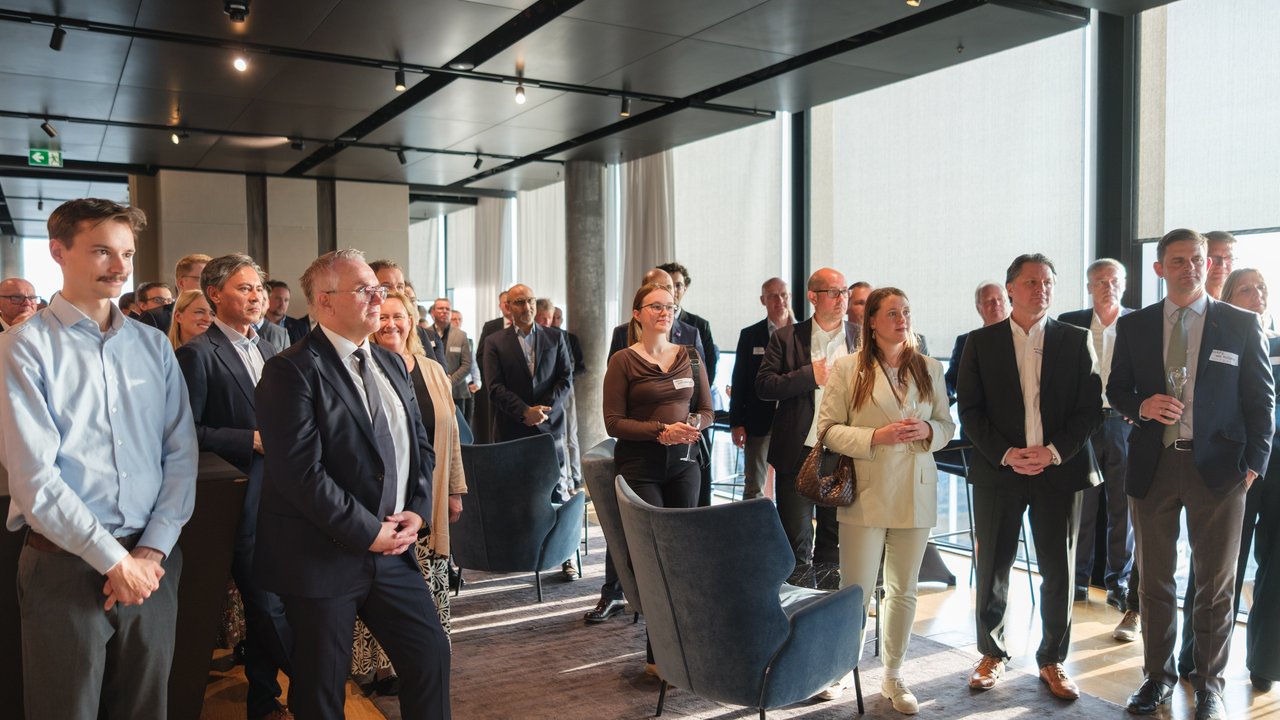 A group of people in business attire gathered in a modern indoor space, some holding drinks and engaged in conversation.