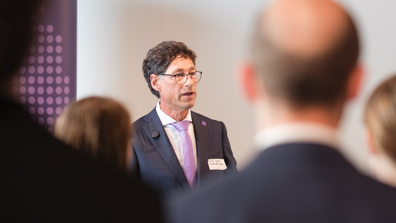 A man in a suit speaks to an audience, partially obscured by attendees in the foreground.