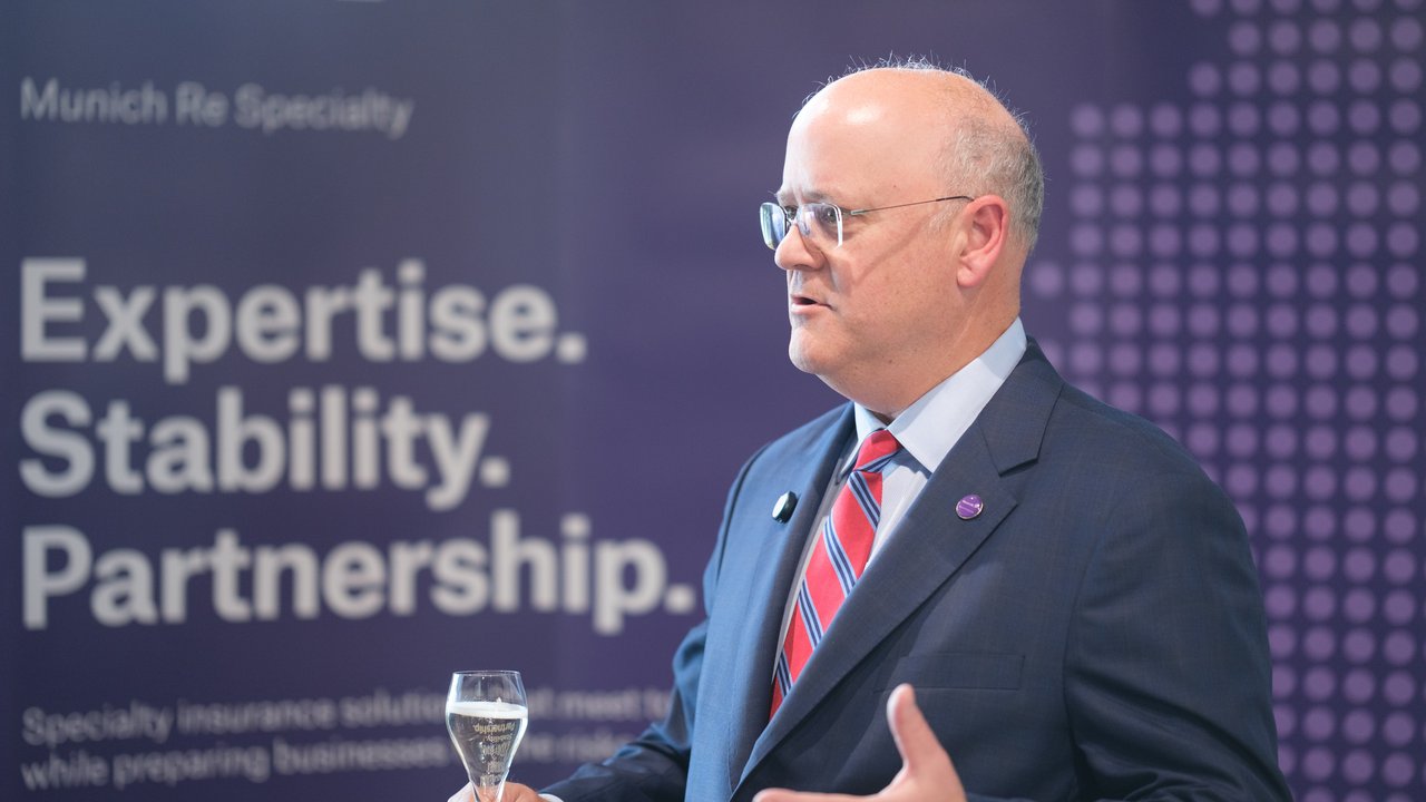 A man in a suit holds a glass while speaking in front of a purple backdrop with text about Munich Re.