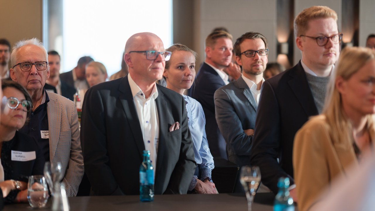 A group of people attentively listening at an event, with drinks on a table in the foreground.