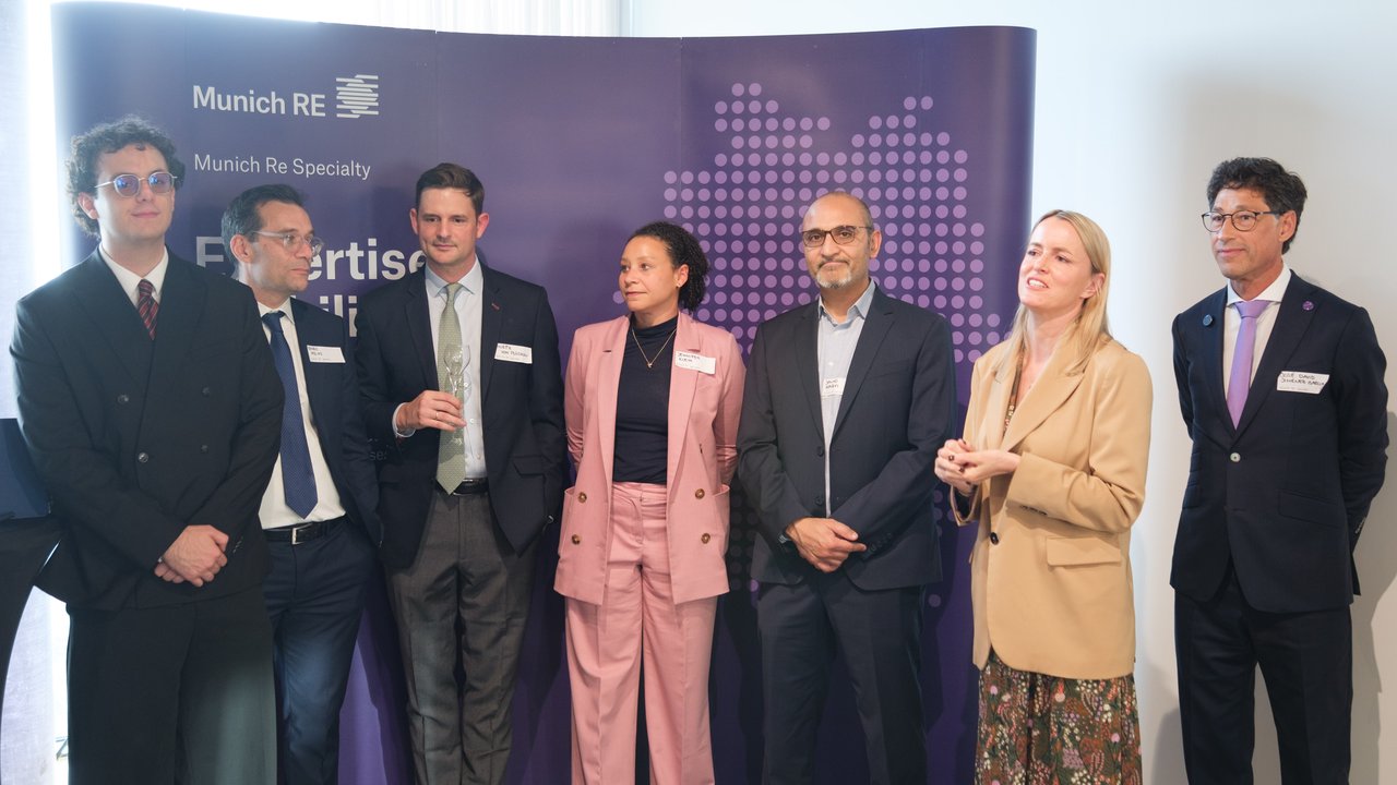 A group of seven professionals stands in front of a purple backdrop with Munich Re branding, engaged in conversation.
