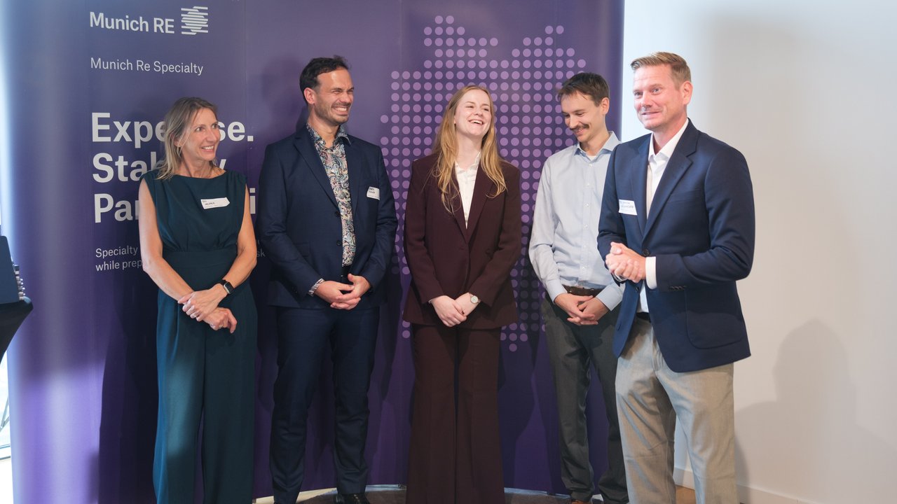 Five people stand in front of a purple backdrop with the Munich Re logo, smiling and engaged in conversation.
