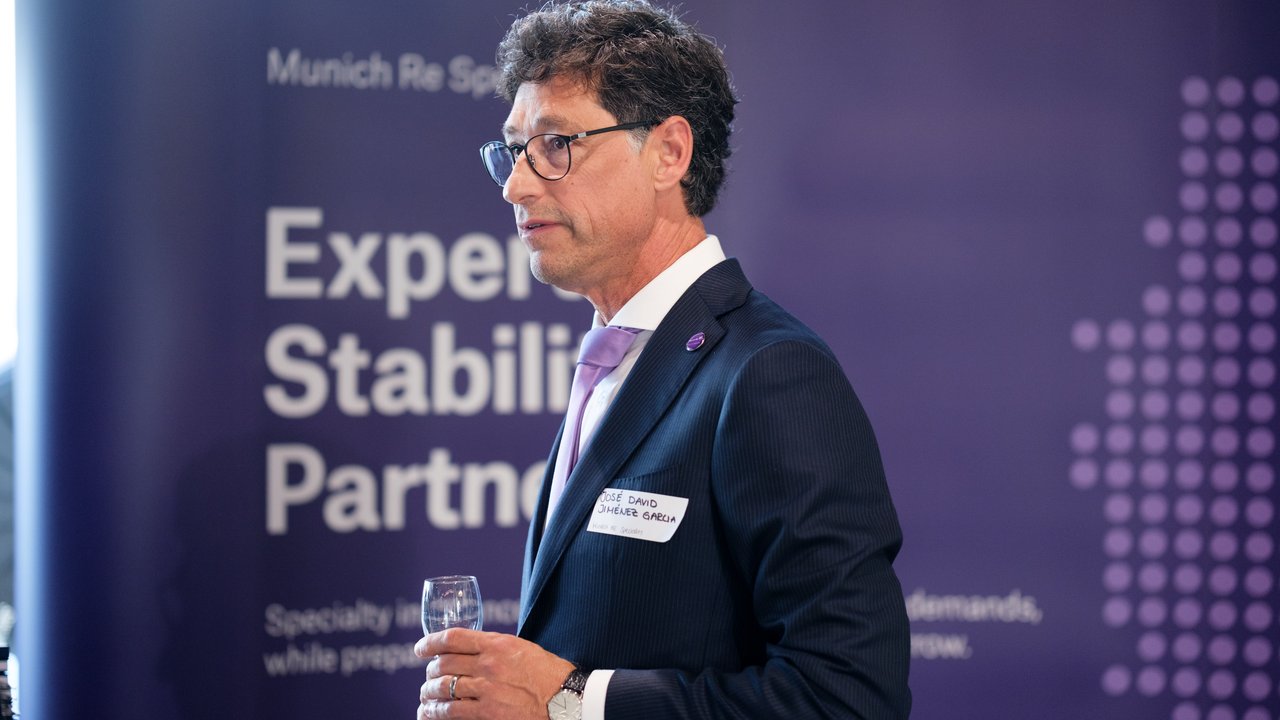 A man in a suit holds a glass, standing in front of a purple backdrop with text about Munich Re.