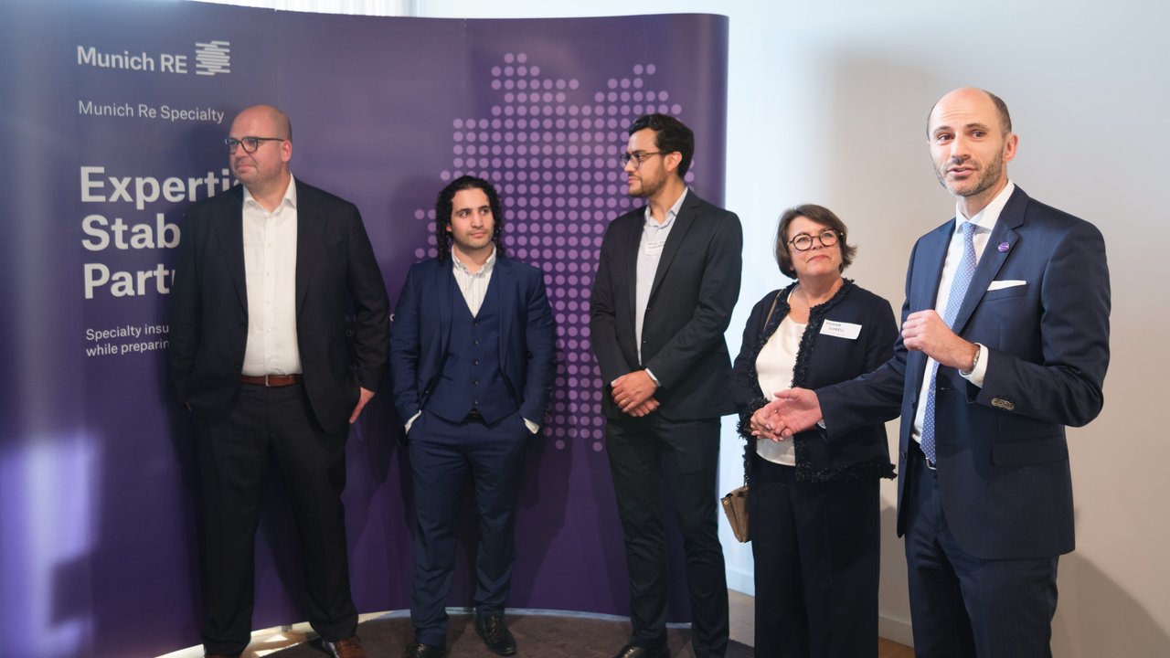 Five people stand in front of a purple backdrop with the Munich RE logo, engaged in conversation.
