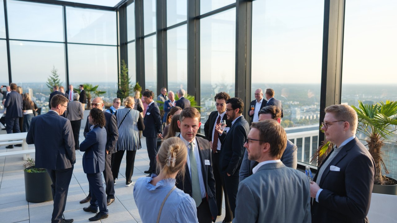 A rooftop event with people in business attire socializing, surrounded by glass walls and city views.