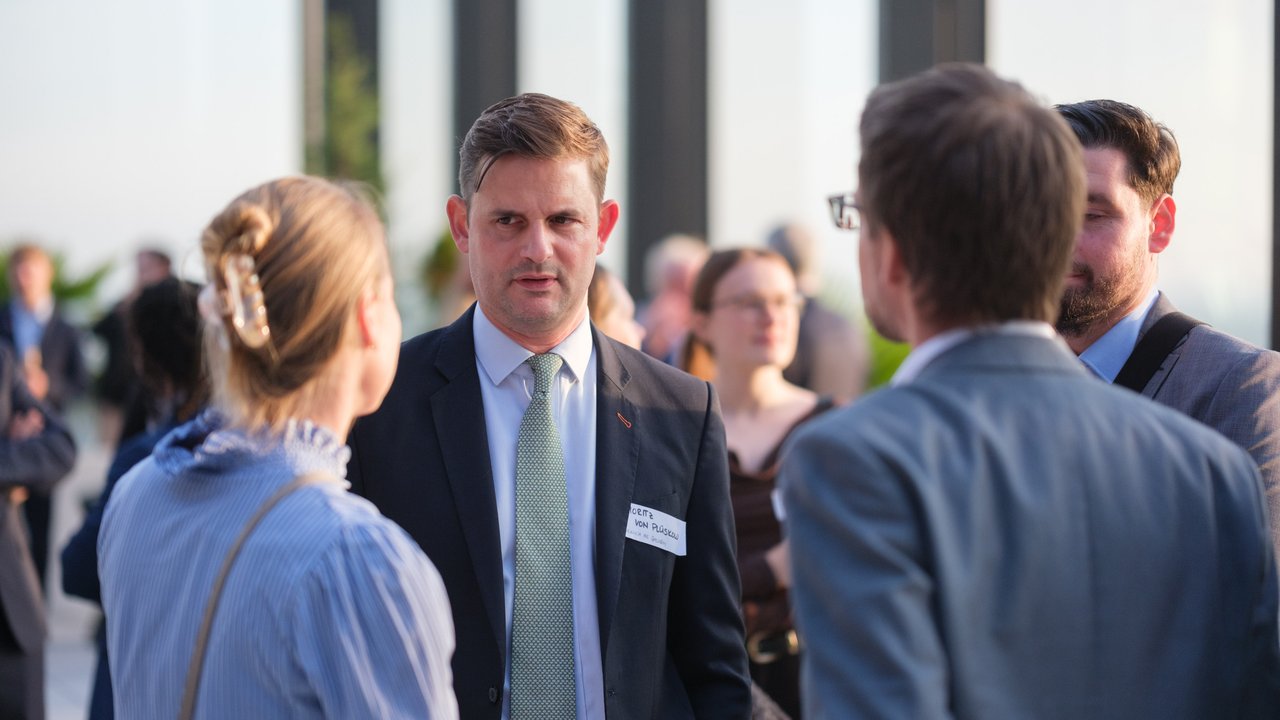 A group of people in business attire engaged in conversation at an outdoor event with a city view in the background.