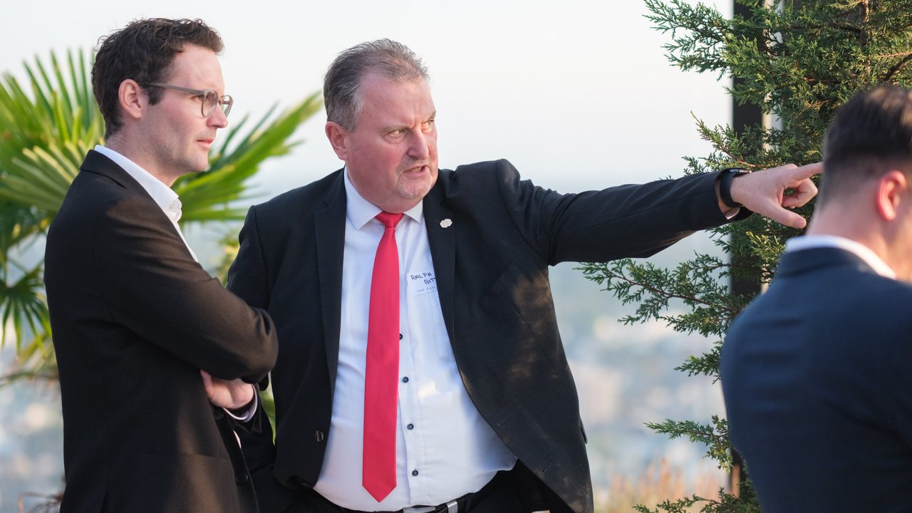 Two men in suits converse outdoors, with one gesturing while the other listens, surrounded by greenery.