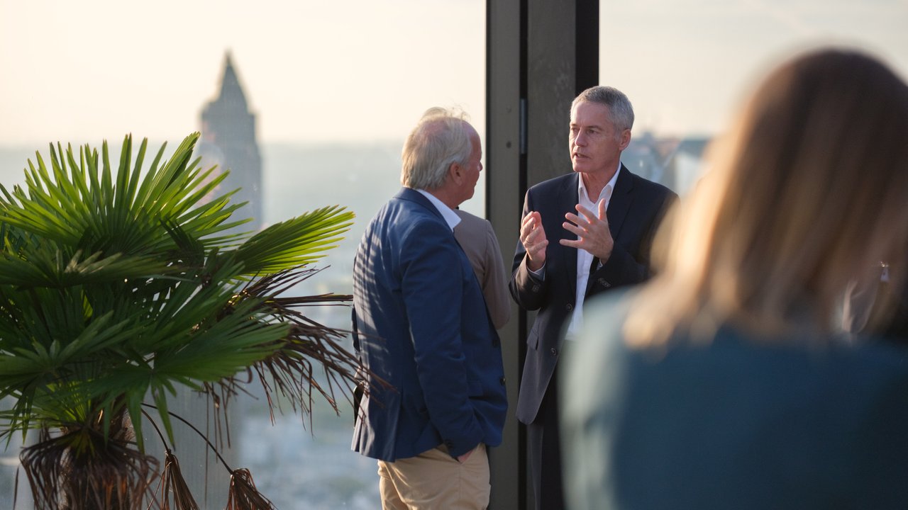 Two men converse in a modern setting with a palm plant and a city skyline visible in the background.