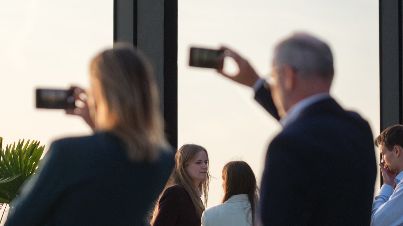 A group of people taking photos with smartphones against a sunset backdrop, with plants in the foreground.