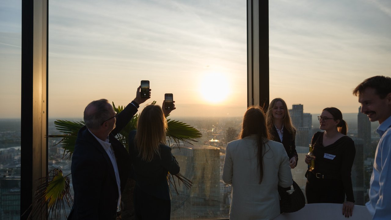 Group of people enjoying a sunset view from a high-rise building, some taking photos with their phones.