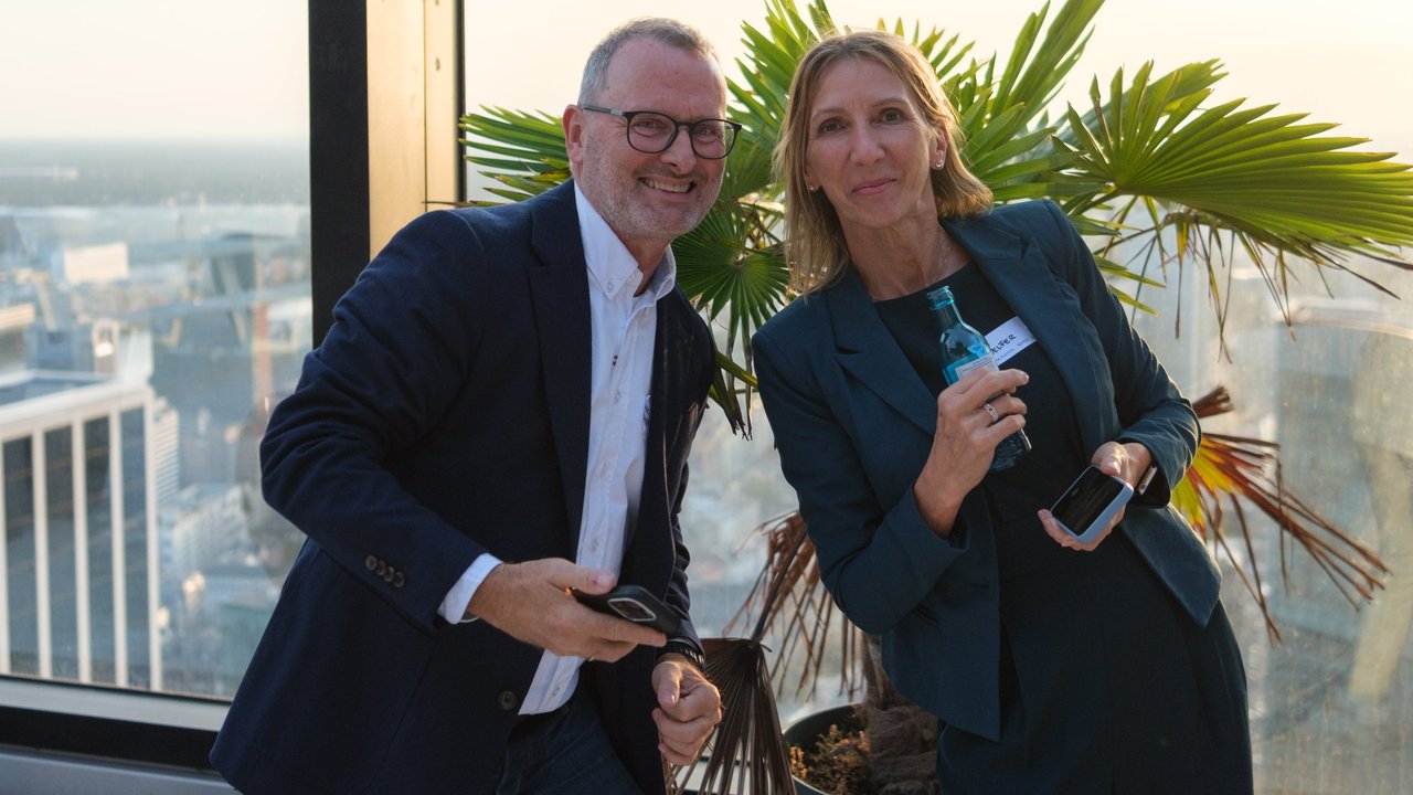 A man and a woman pose together near a large window with a city view, holding smartphones and smiling.