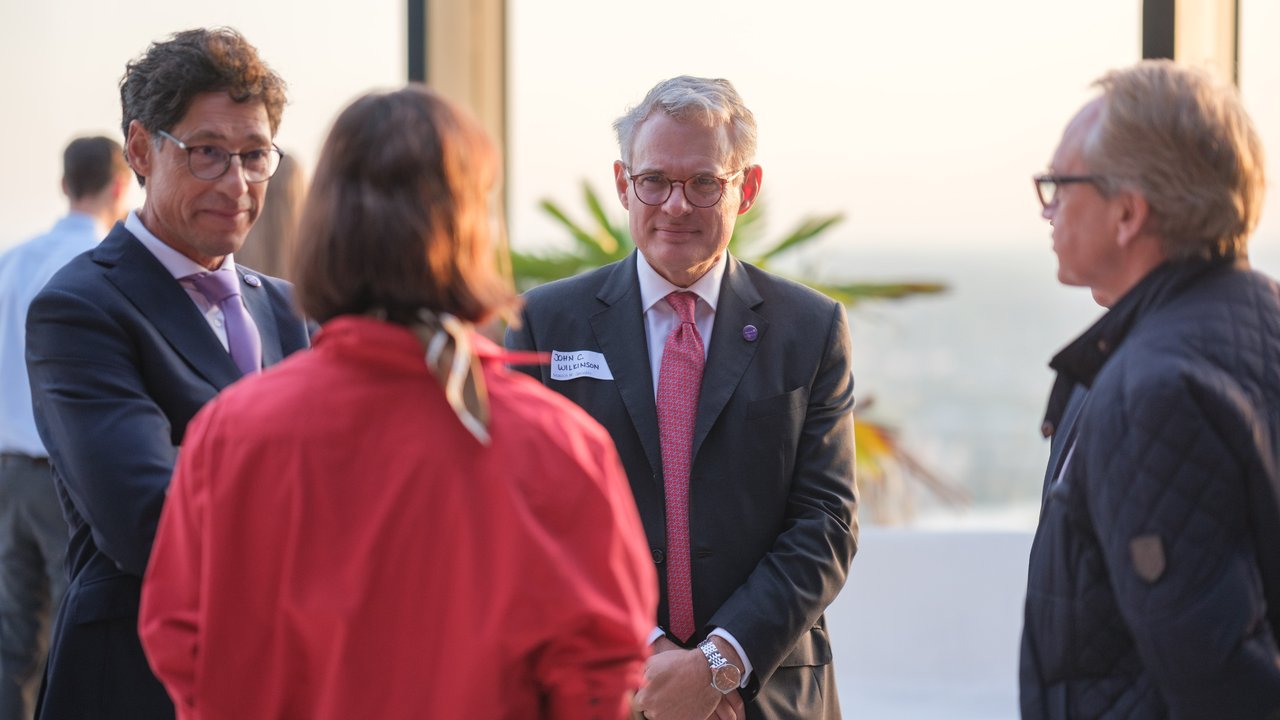 Four people in business attire engage in conversation at a social event with a city view in the background.
