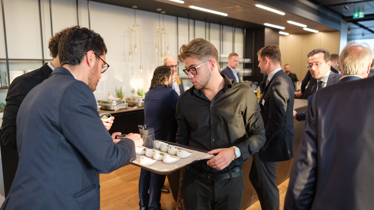 A group of people in business attire socializing at an event, with one person serving food on a tray.
