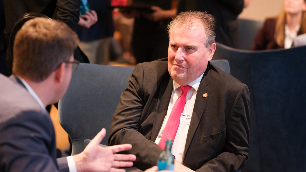 Two men are seated in a conference setting, one speaking animatedly while the other listens attentively.