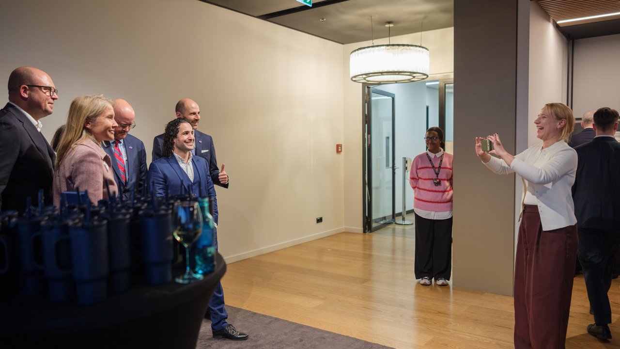 A group of six people pose for a photo in a modern indoor setting, with one person taking the picture.