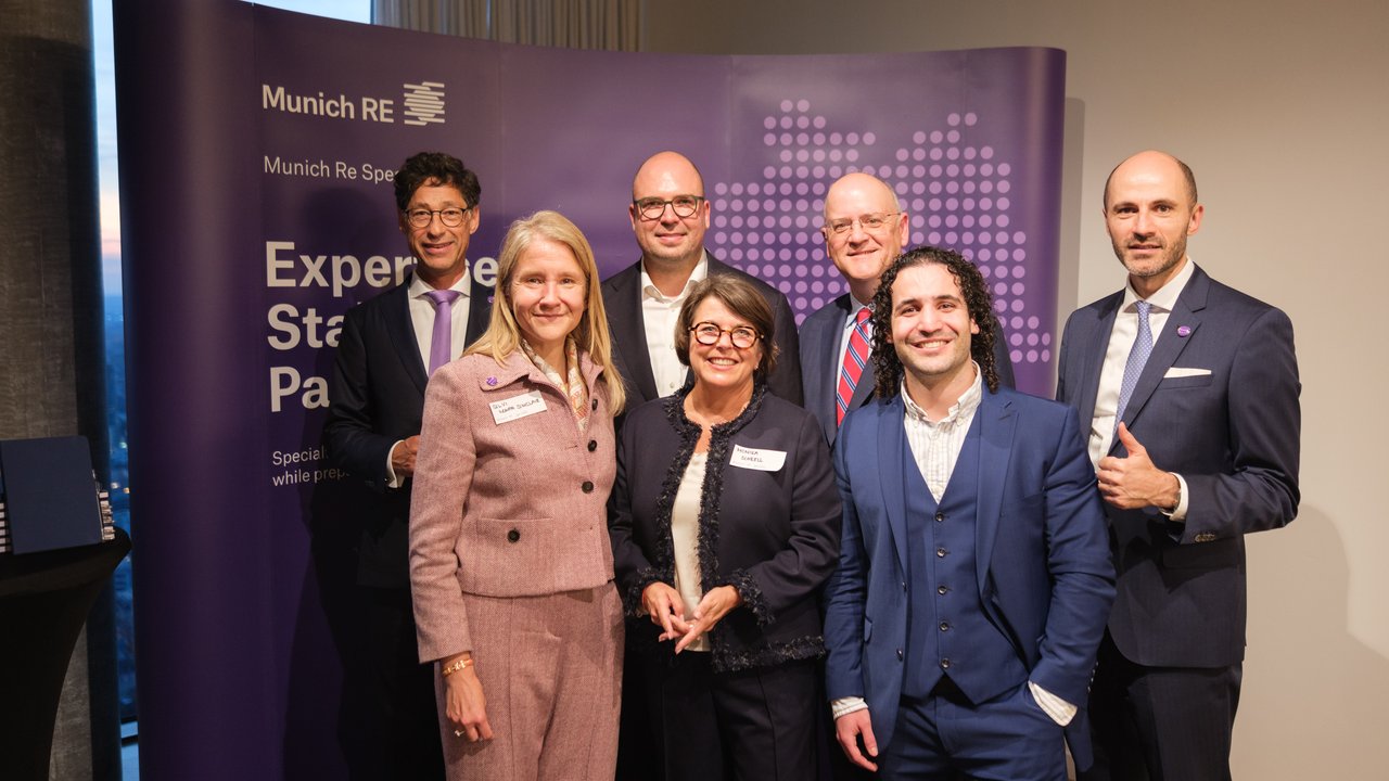 A group of seven professionals poses in front of a purple Munich Re banner at an event.