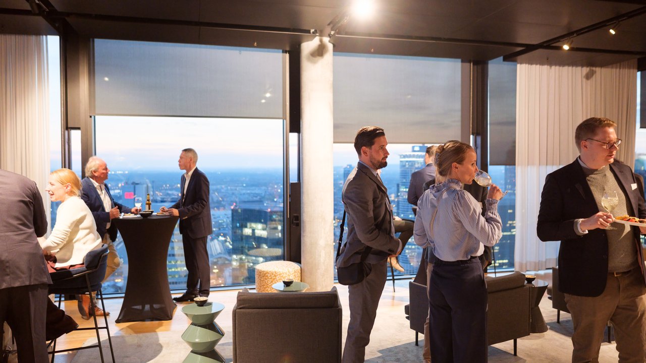 A group of people socializing in a modern room with large windows overlooking a city skyline at dusk.