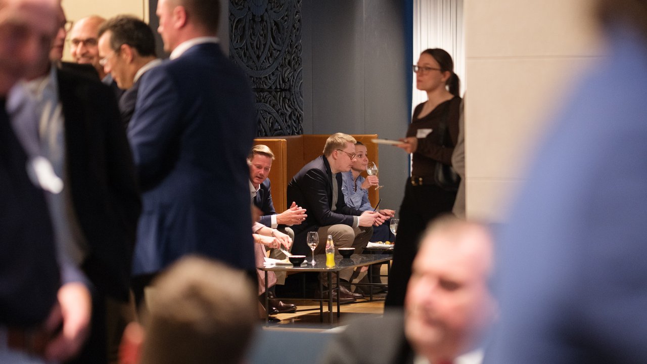 A group of people mingling at an event, with some seated and others standing, in a modern indoor setting.