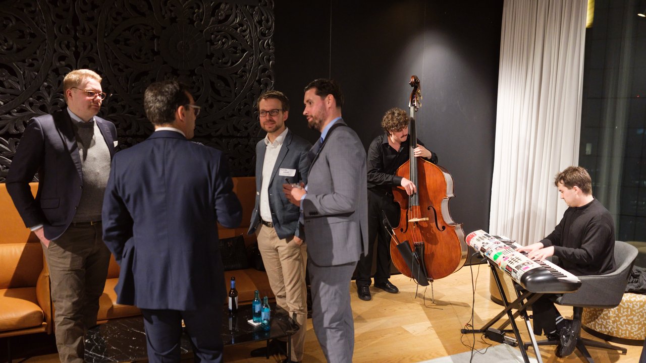 A group of men converse in a lounge while musicians play a keyboard and double bass in the background.