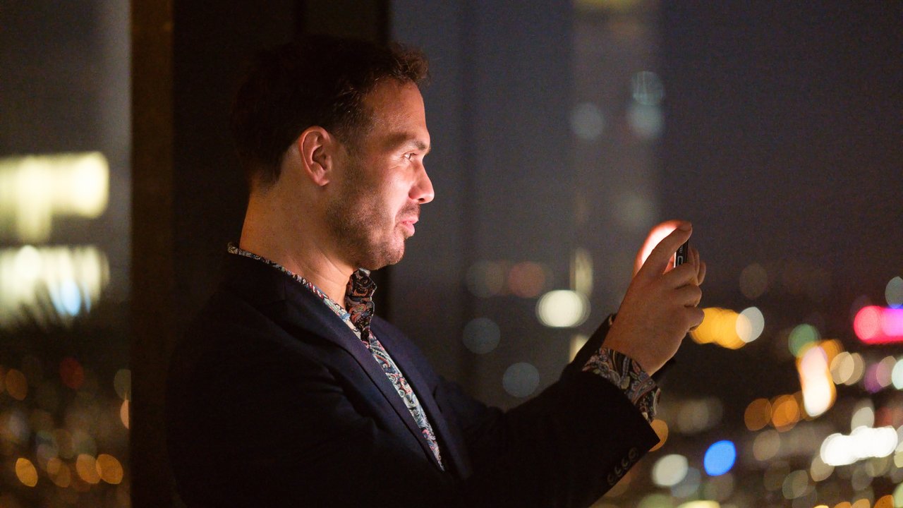 A man in a dark suit takes a photo with a smartphone against a blurred city night backdrop.