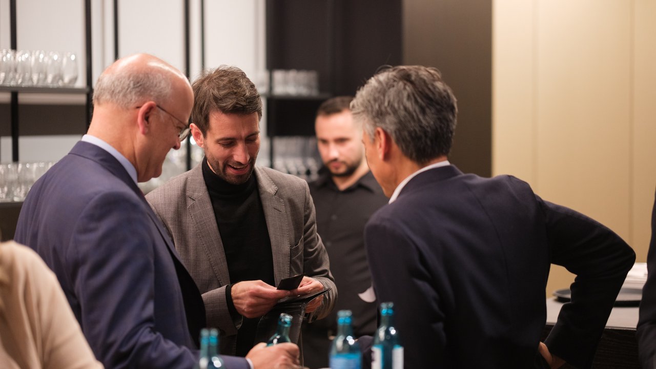Four men engage in conversation at a bar, with drinks and glasses visible in the background.