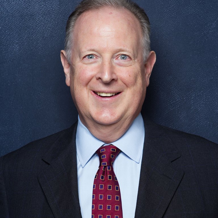 A man in a dark suit and patterned tie stands against a textured blue background.