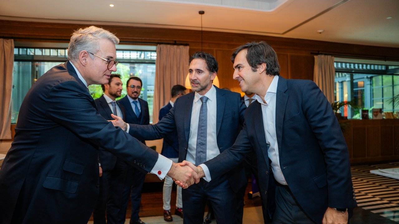 Two men shake hands in a well-lit room with wooden decor and large windows, surrounded by other individuals.