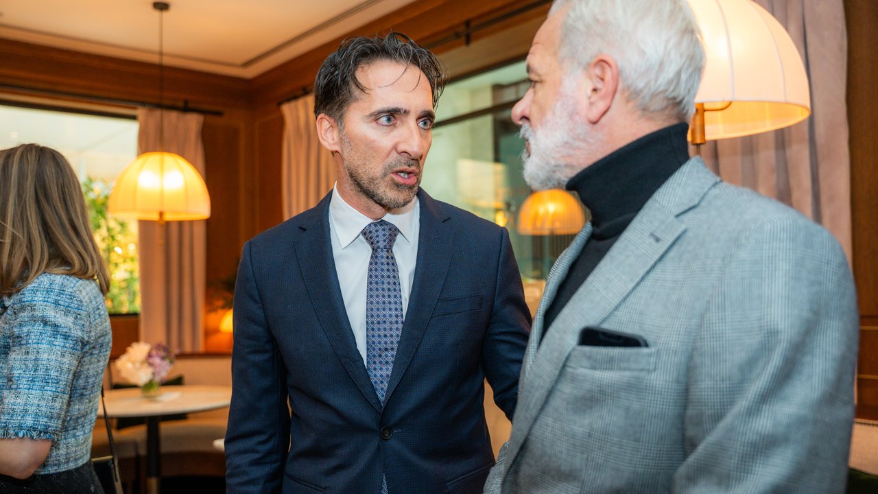Two men in formal attire converse in a stylish indoor setting with soft lighting and decorative lamps.