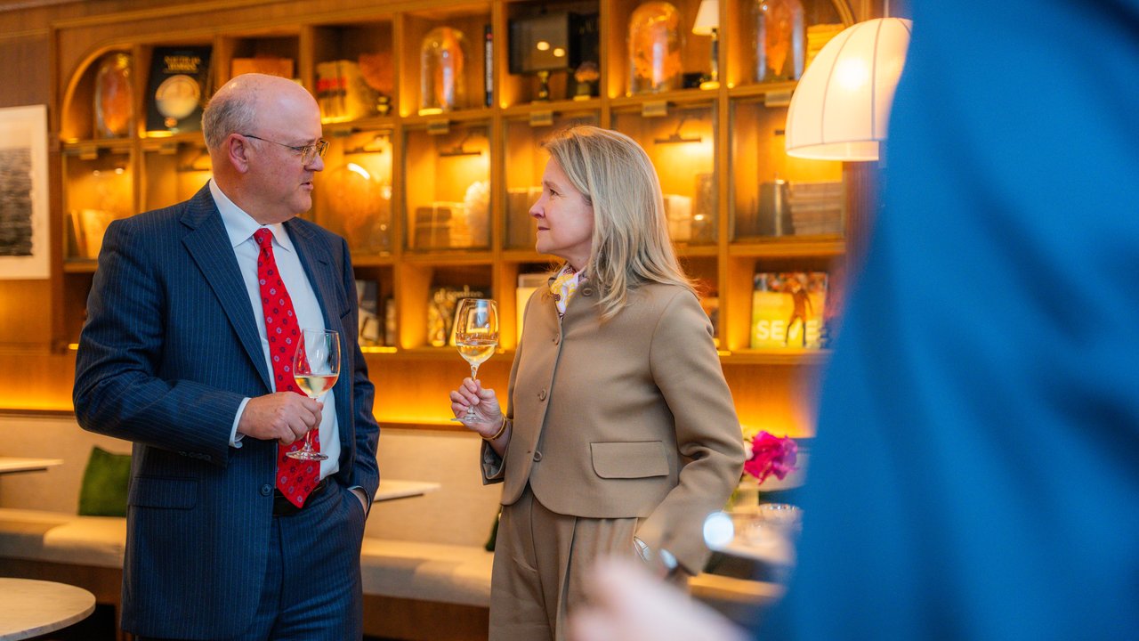 A man in a suit and a woman in a beige outfit hold wine glasses in a stylish bar setting.