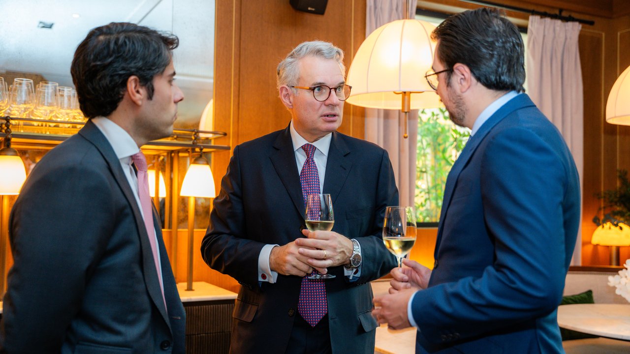 Three men in formal attire are engaged in conversation, holding glasses of wine in a well-lit indoor setting.