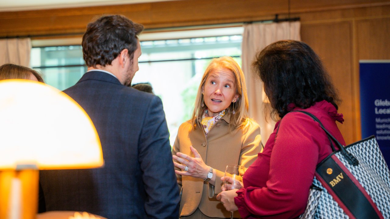 A group of four people engaged in conversation at a social event, with drinks in hand and a lamp in the foreground.