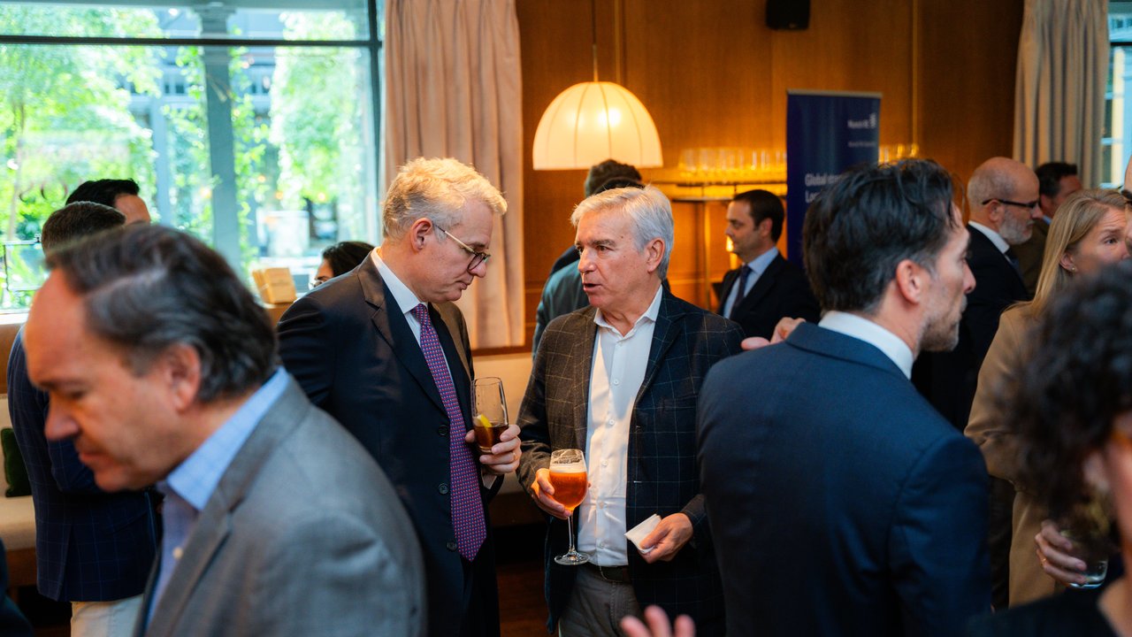 A group of people socializing indoors, holding drinks, with large windows and a modern decor in the background.