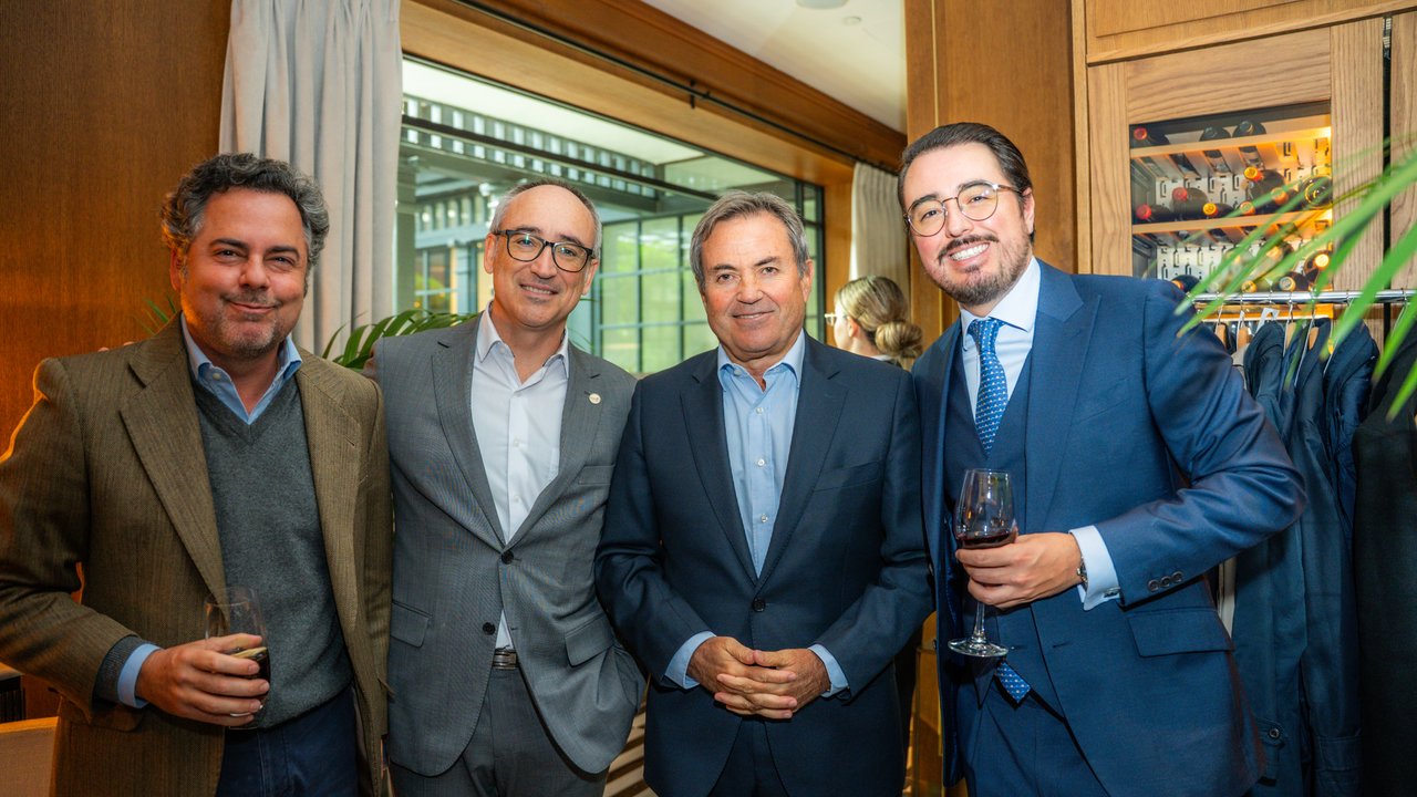 Four men stand together in a well-lit room, holding drinks, with a wine rack and plants in the background.