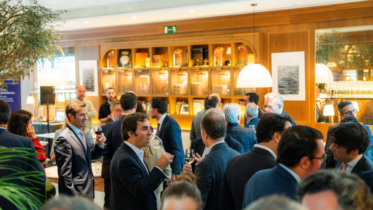 A group of people in formal attire socializing in a well-lit indoor space with shelves and decorative items.