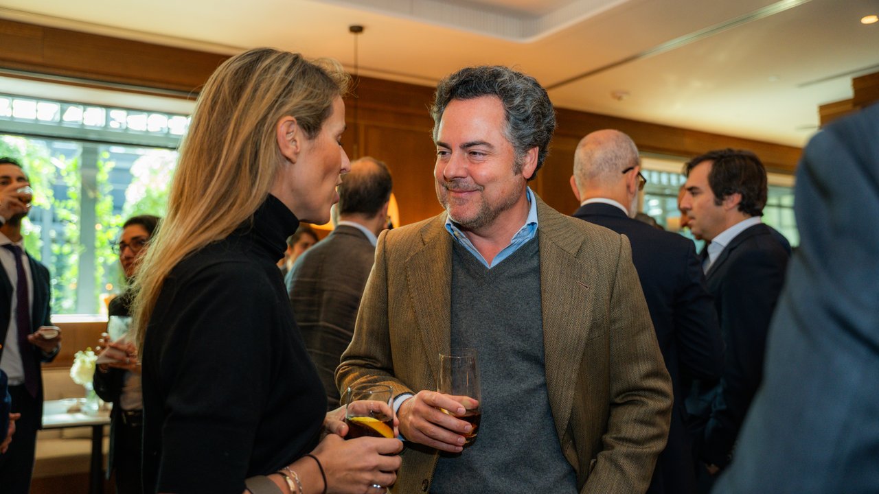 A man in a blazer converses with a woman in a black top at a social gathering, surrounded by other attendees.