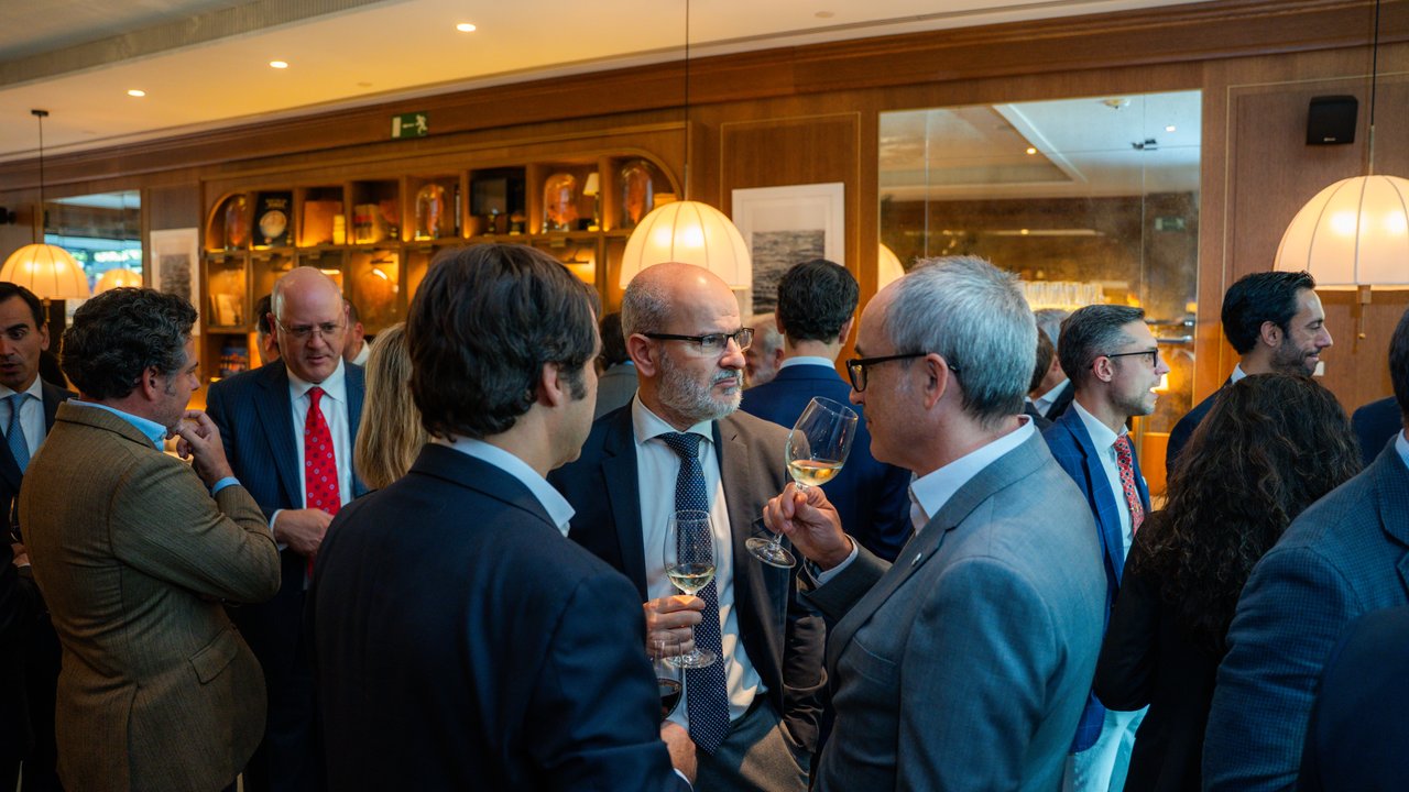 A group of people in formal attire socializing in a well-lit indoor setting with drinks in hand.