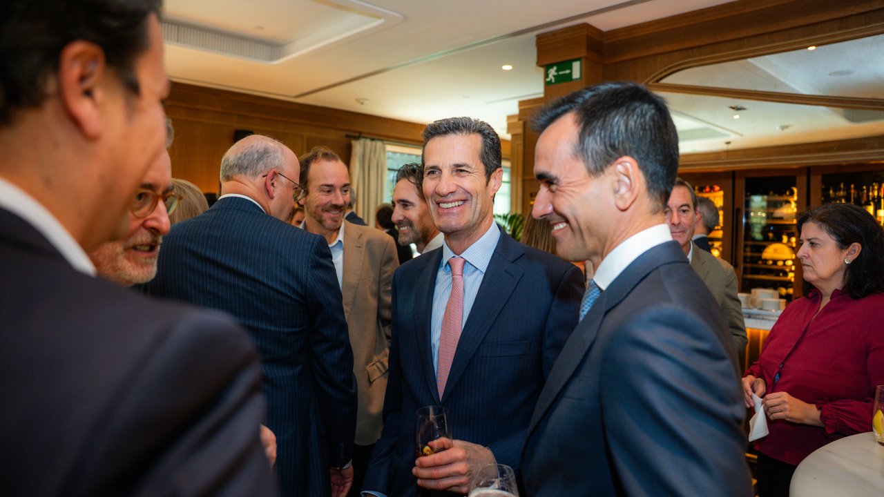 A group of people in formal attire socializing at a gathering, with drinks in hand and a bar in the background.