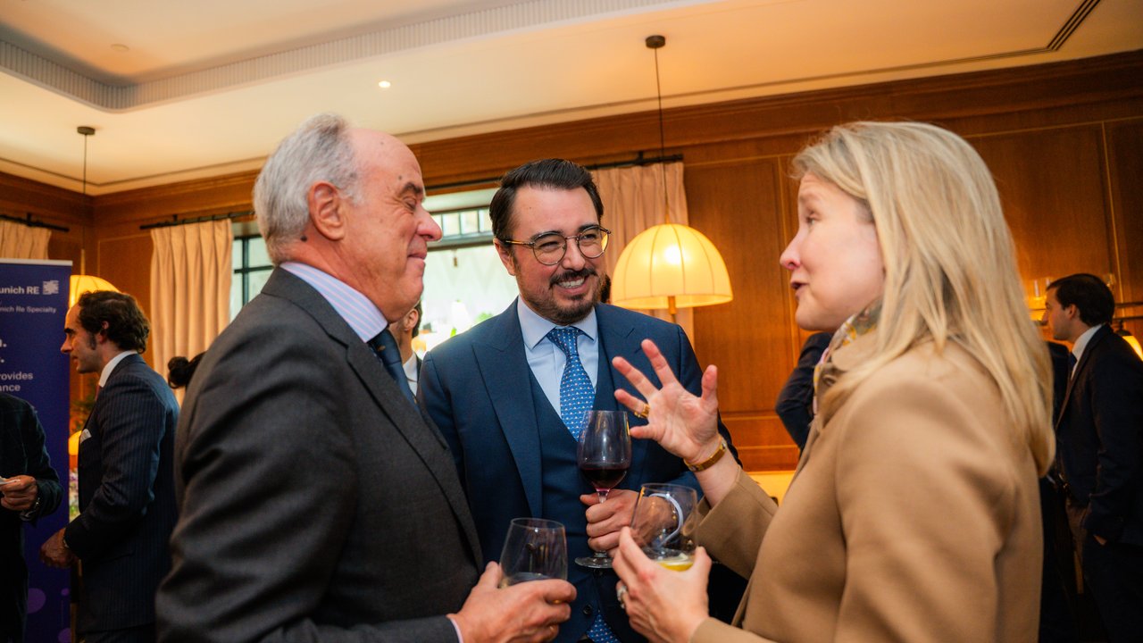 Three people engaged in conversation at a social event, holding drinks in a well-lit indoor setting.