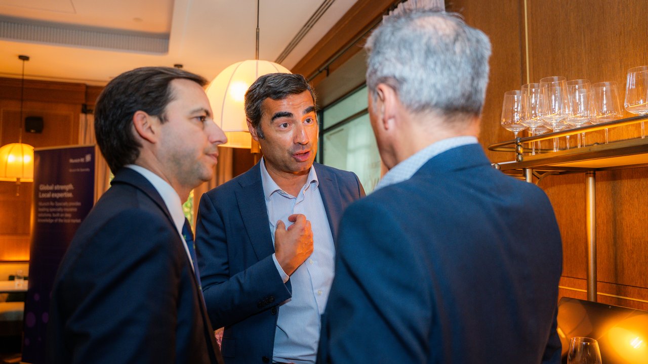 Three men in suits are engaged in conversation in a well-lit indoor setting with glasses on a shelf in the background.