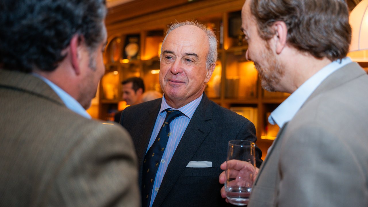 Three men in conversation at a social gathering, with a wooden shelf filled with items in the background.