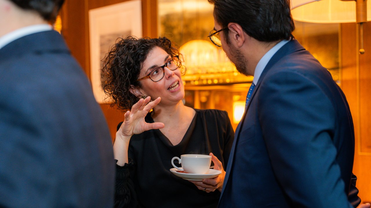 A woman in a black top holds a cup and saucer while conversing with a man in a suit.