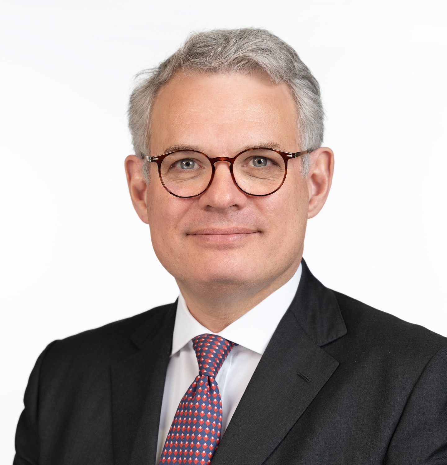 A man in a dark suit and patterned tie stands against a white background, face obscured.