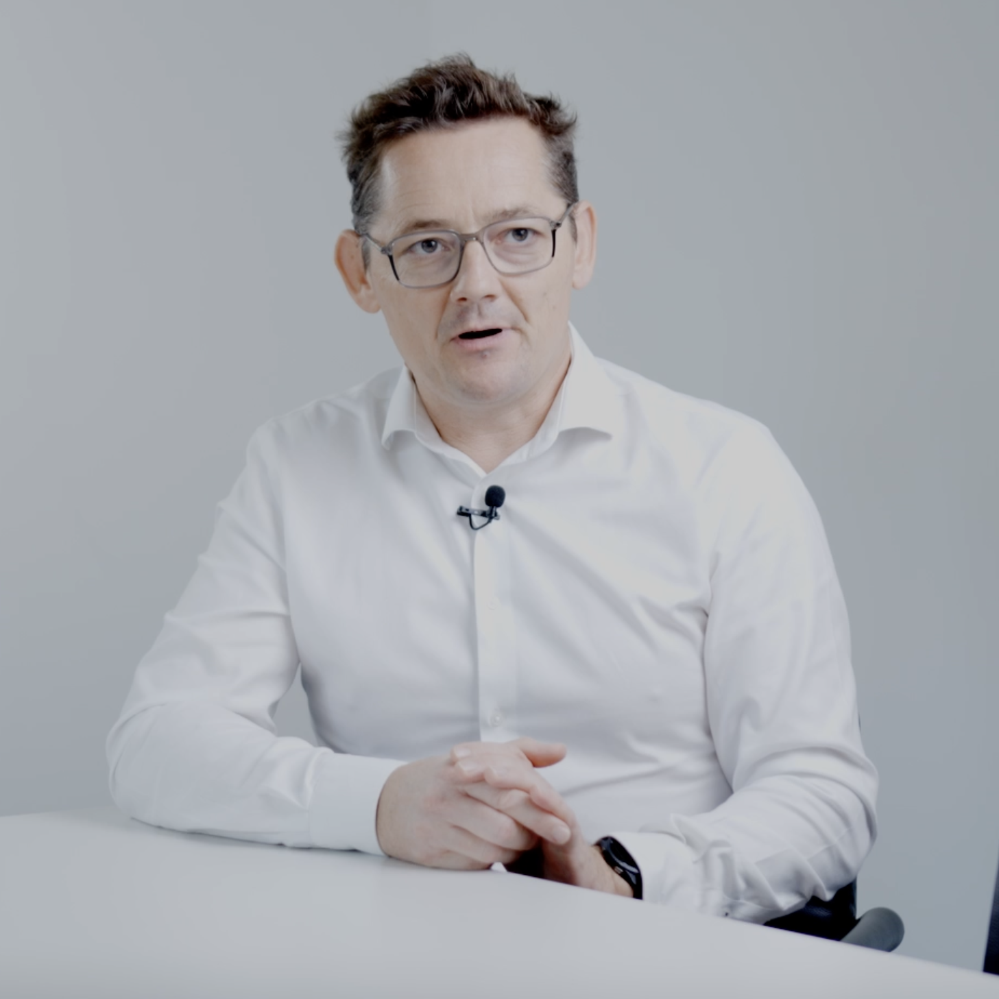A person in a white shirt sits at a table, hands clasped, with a microphone attached.